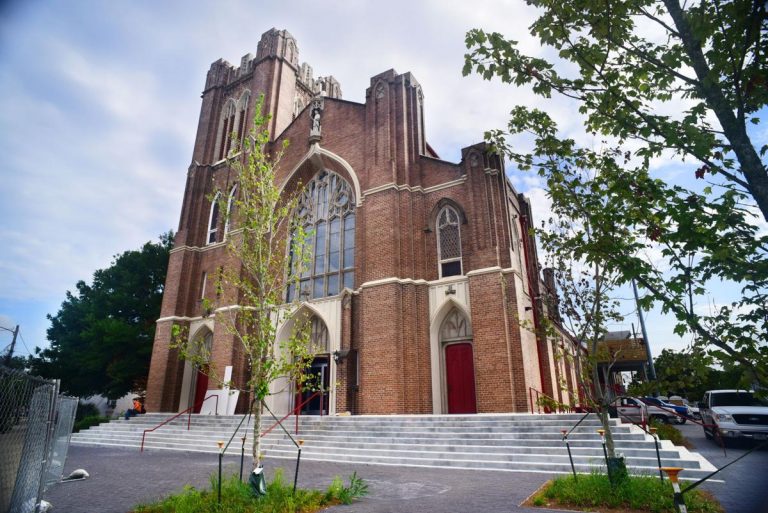 An old church building, made of reddish brick with white accents and red doors. One side has a tower with turrets at the top. The parking area beside it is dotted with young green trees.