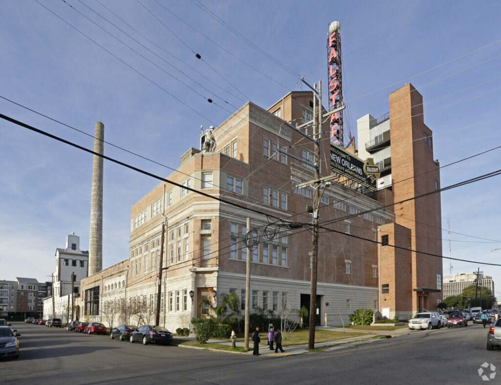 A large building made of tan and white brick, with a tower rising above it that reads "Falstaff" in large red letters. Cars are parked on the road alongside its tidy lawn and sidewalks.