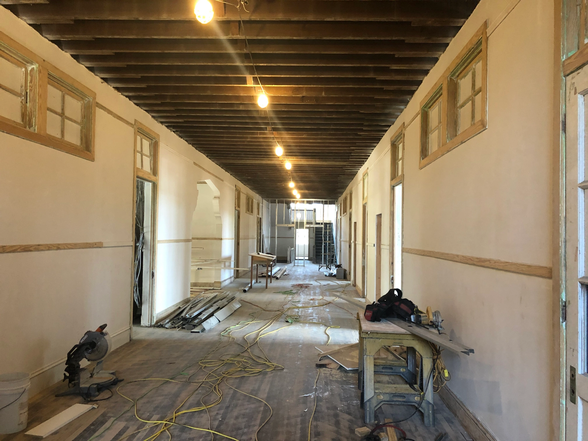 An interior hallway in the Tate, Etienne, and Prevost Center during during lead and asbestos abatement in 2020. The ceiling rafters are visible with light bulbs strung along them, and the floor is covered in extension cords, lumber, and tools.
