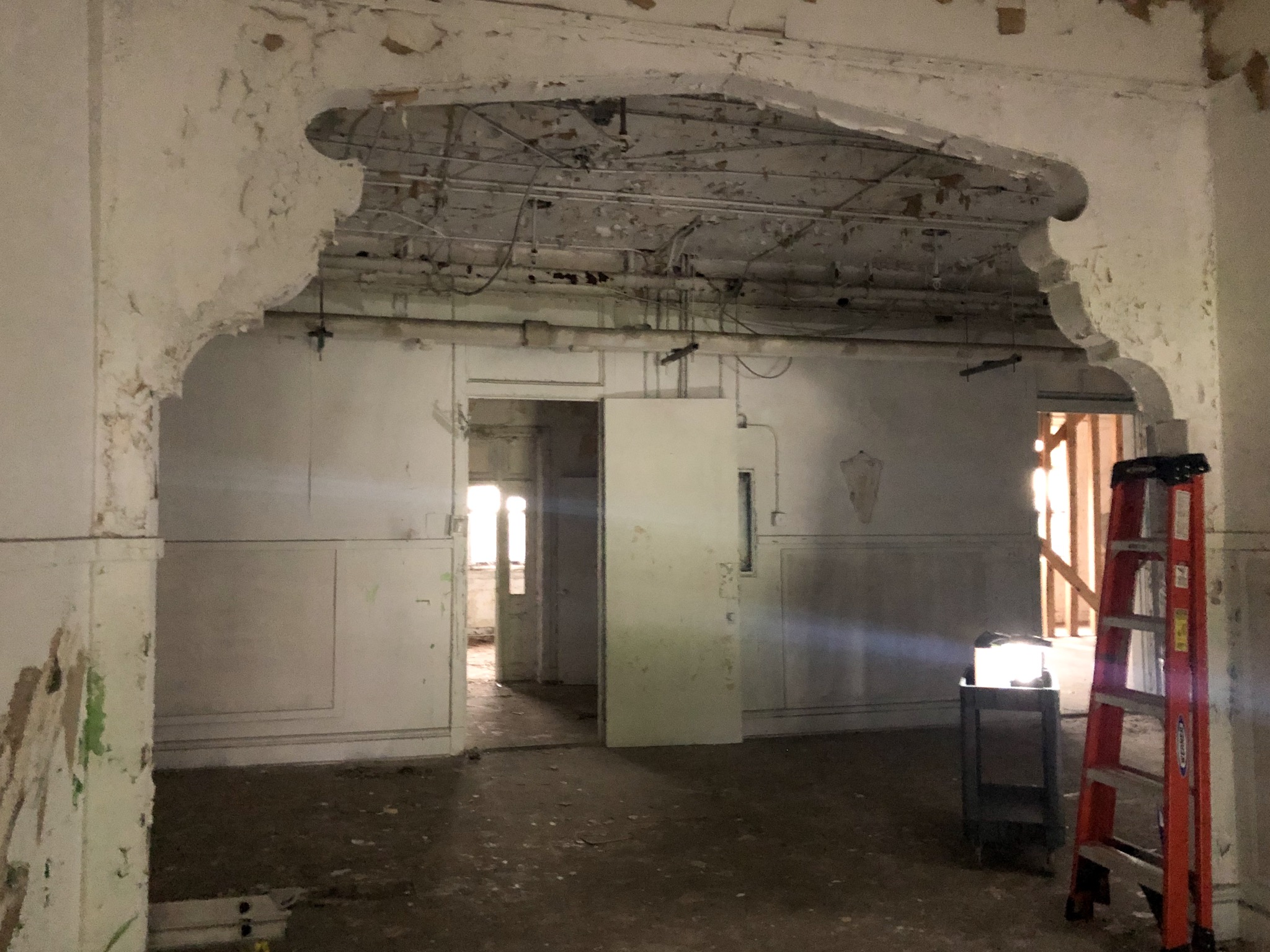 The interior of the Tate, Etienne, and Prevost Center during the Phase II environmental assessment. The photo depits a room with a ladder leaning against the wall, pipes visible in the ceiling and a wall removed in the background. A decorative archway frames the entrance.
