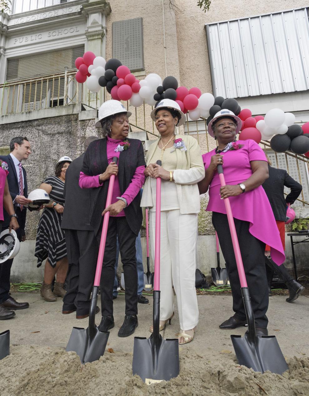 Three Black women, Gail Etienne, Tessie Prevost, and Leona Tate, stand together wearing hard hats and holding shovels with pink handles. Etienne and Tate are wearing hot pink and black, and Prevost is wearing offwhite. They wear hot pink corsages and there are pink, white, and black balloons behind them, tied to the stairway railing of the former McDonogh 19 school that now bears their names. Photo Credit: Nola.com
